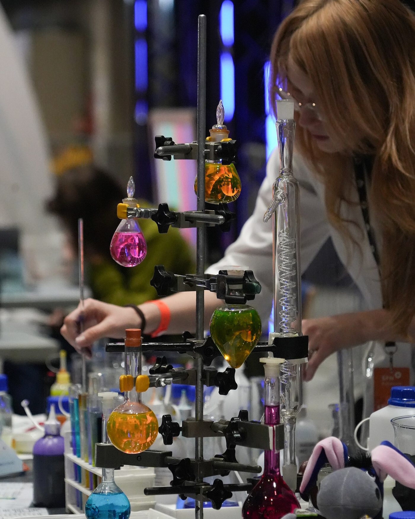 A researcher at a science festival pours a coloured liquid between glass vessels at a chemistry demonstration stand.