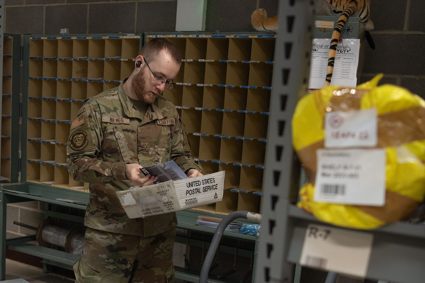 A postal clerk in uniform sorts a handful of inbound letters in front of rows of pigeonhole shelves.