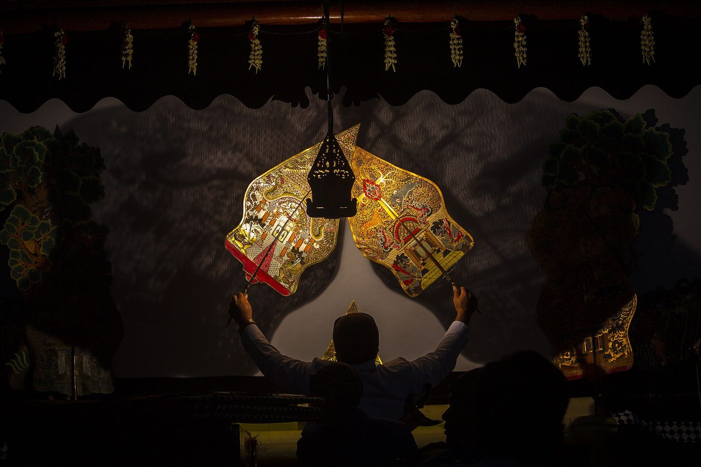 A wayang kulit performance seen from behind the screen: an ornate leather shadow puppet lit against a white screen, the puppeteer’s silhouette below.