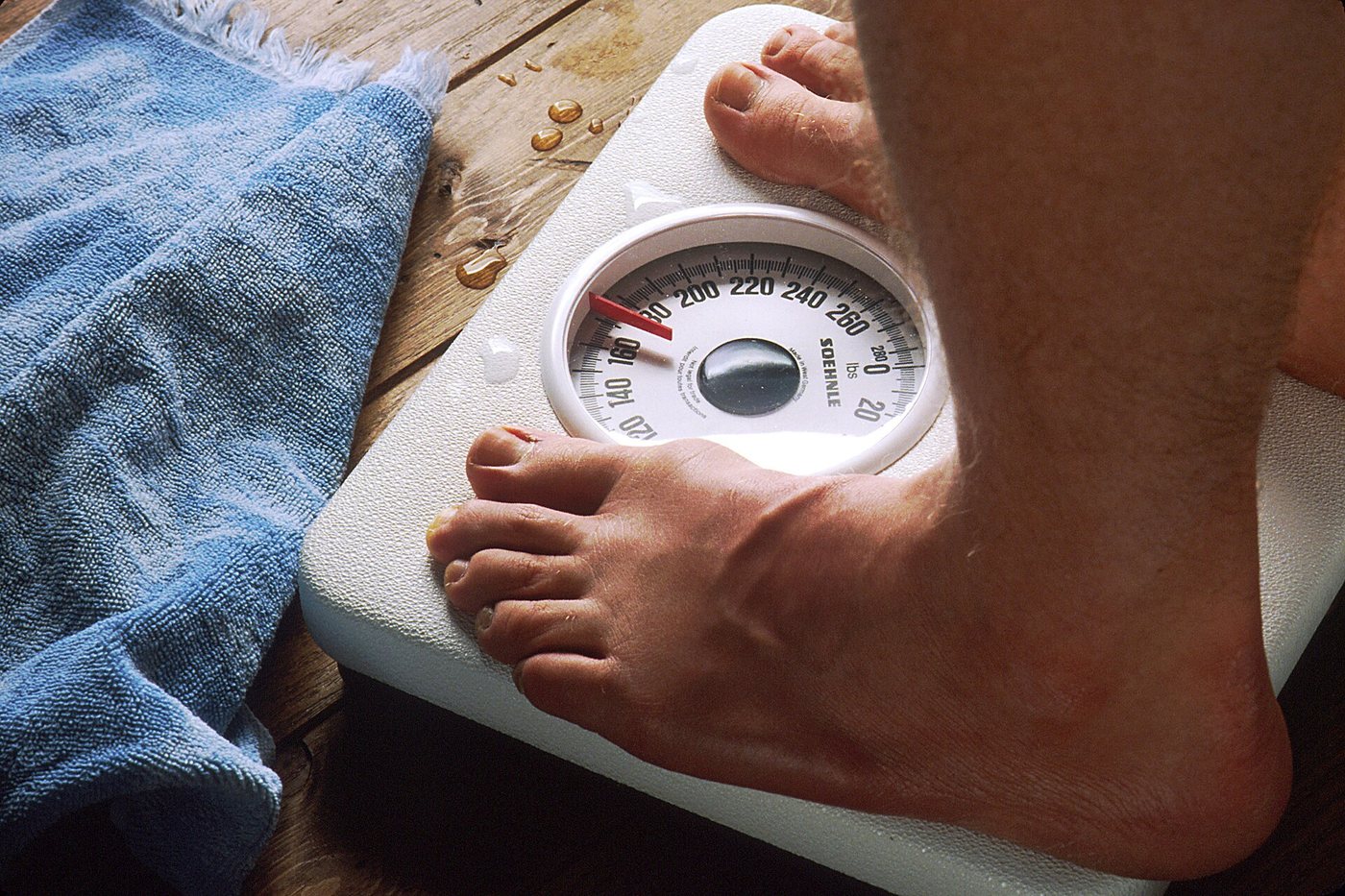 Des pieds nus sur un pèse-personne analogique à cadran rond, à côté d’une serviette bleue, sur un plancher de bois.