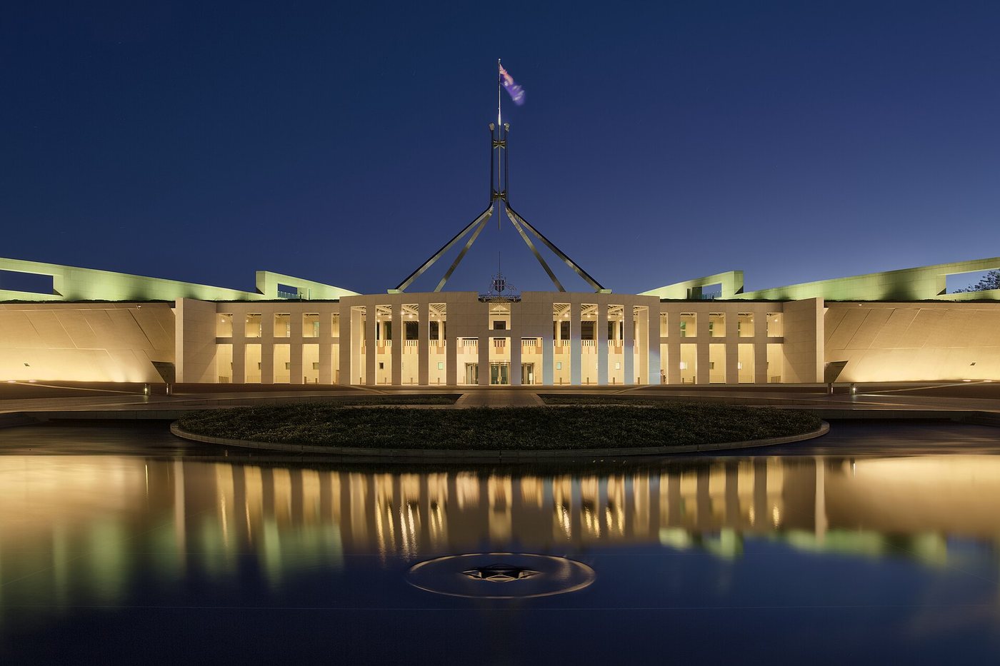 Le Parlement de Canberra, en Australie, au crépuscule, avec son grand mât et sa façade éclairée qui se reflètent dans le bassin du parvis.