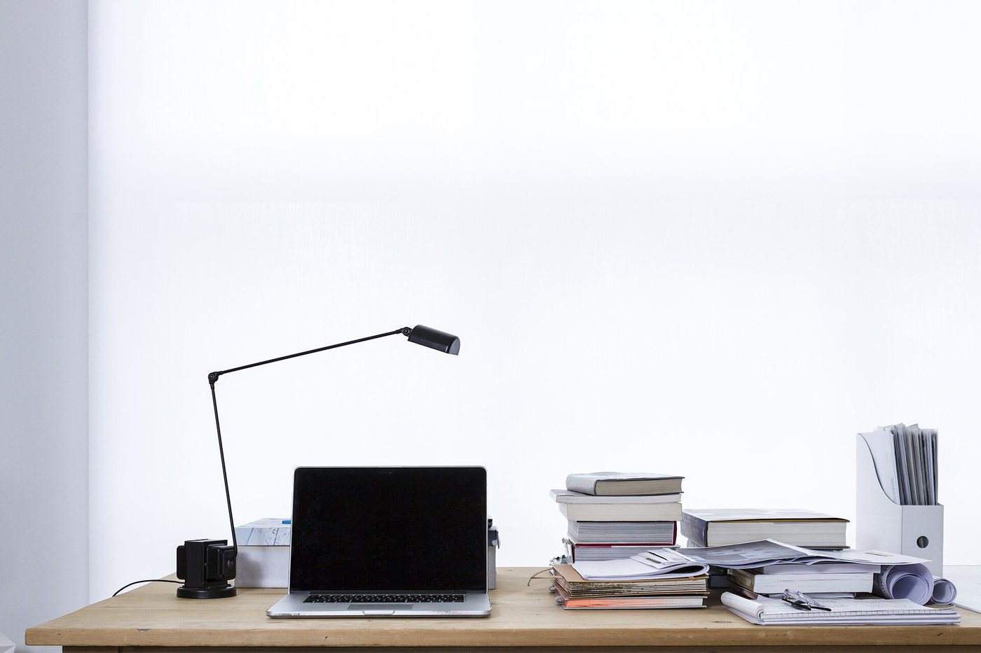 Un ordinateur portable ouvert sur un bureau en bois clair, à côté d’une lampe et de piles de livres, devant un mur blanc.