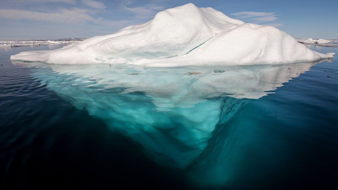 Un iceberg dans une eau arctique calme et claire, dont la partie immergée, bien plus grande, est visible sous la surface.