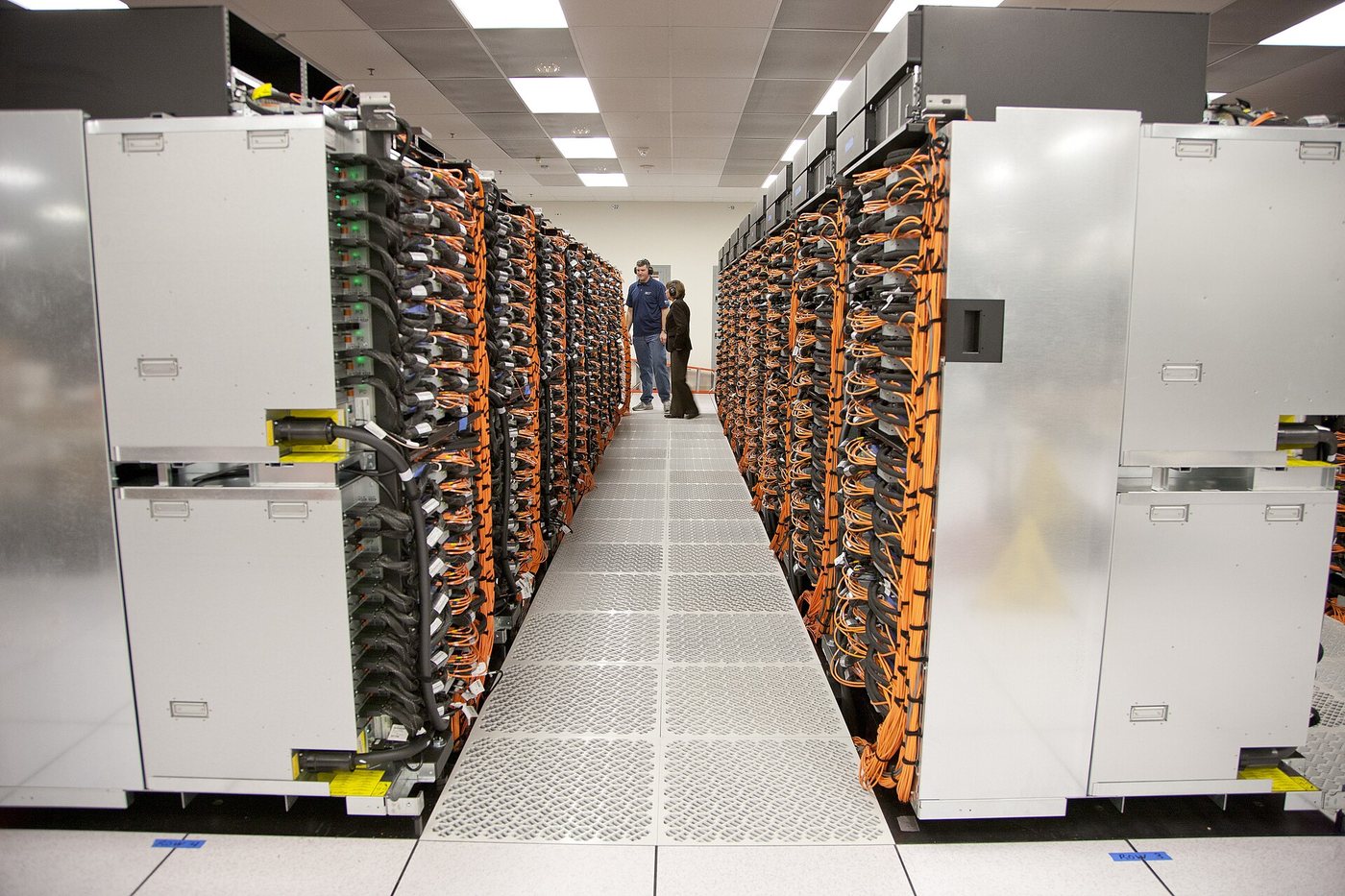 Une longue allée entre les armoires du superordinateur Sequoia au Lawrence Livermore National Laboratory, avec des câbles orange et deux petites silhouettes au fond.